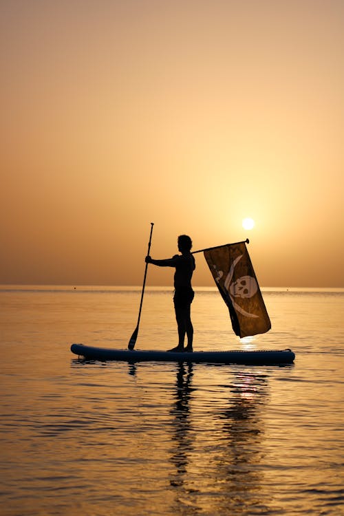 Silhouette of a Man Standing on a Surfboard during Sunset