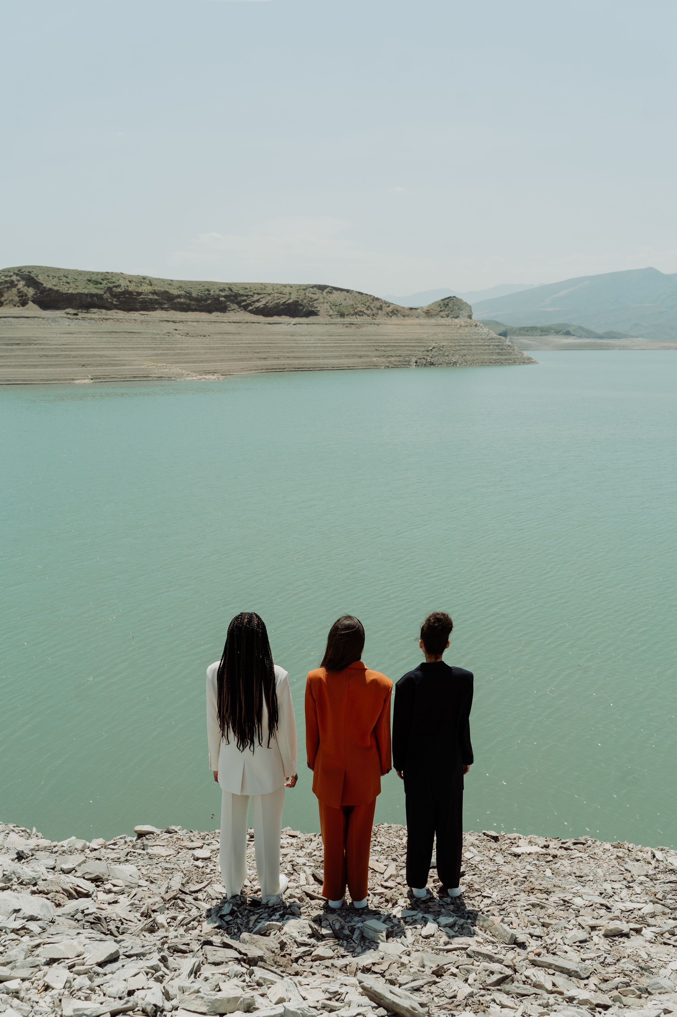 Three stylishly dressed people stand in suits facing a serene lake view.