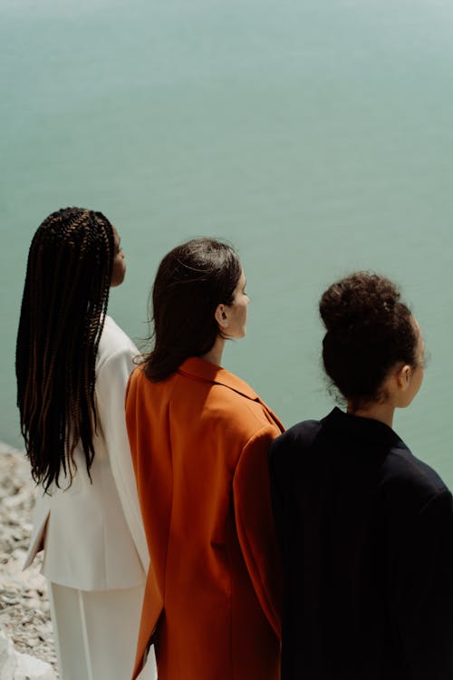 Back View of Women in Suits in Different Colors Standing near a Body of Water 