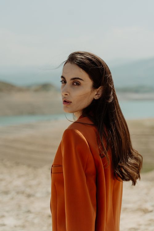 Free Young woman in an orange coat standing by the beach with a thoughtful expression. Stock Photo