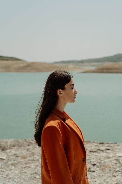 Free Side profile of a young woman in an orange jacket standing by a calm lake under a clear blue sky. Stock Photo