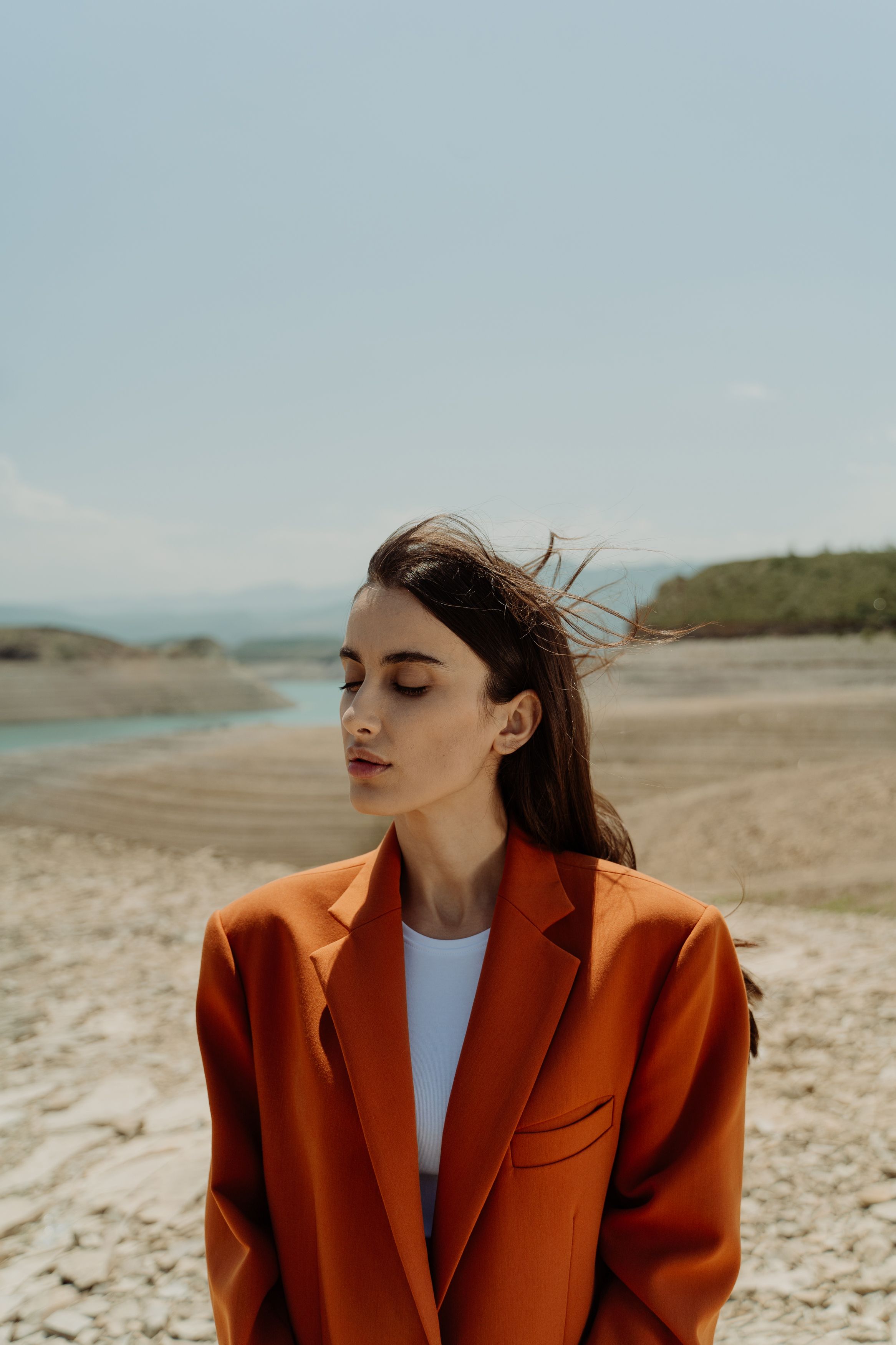 Stylish woman in orange blazer poses under sunlight in a desert landscape.