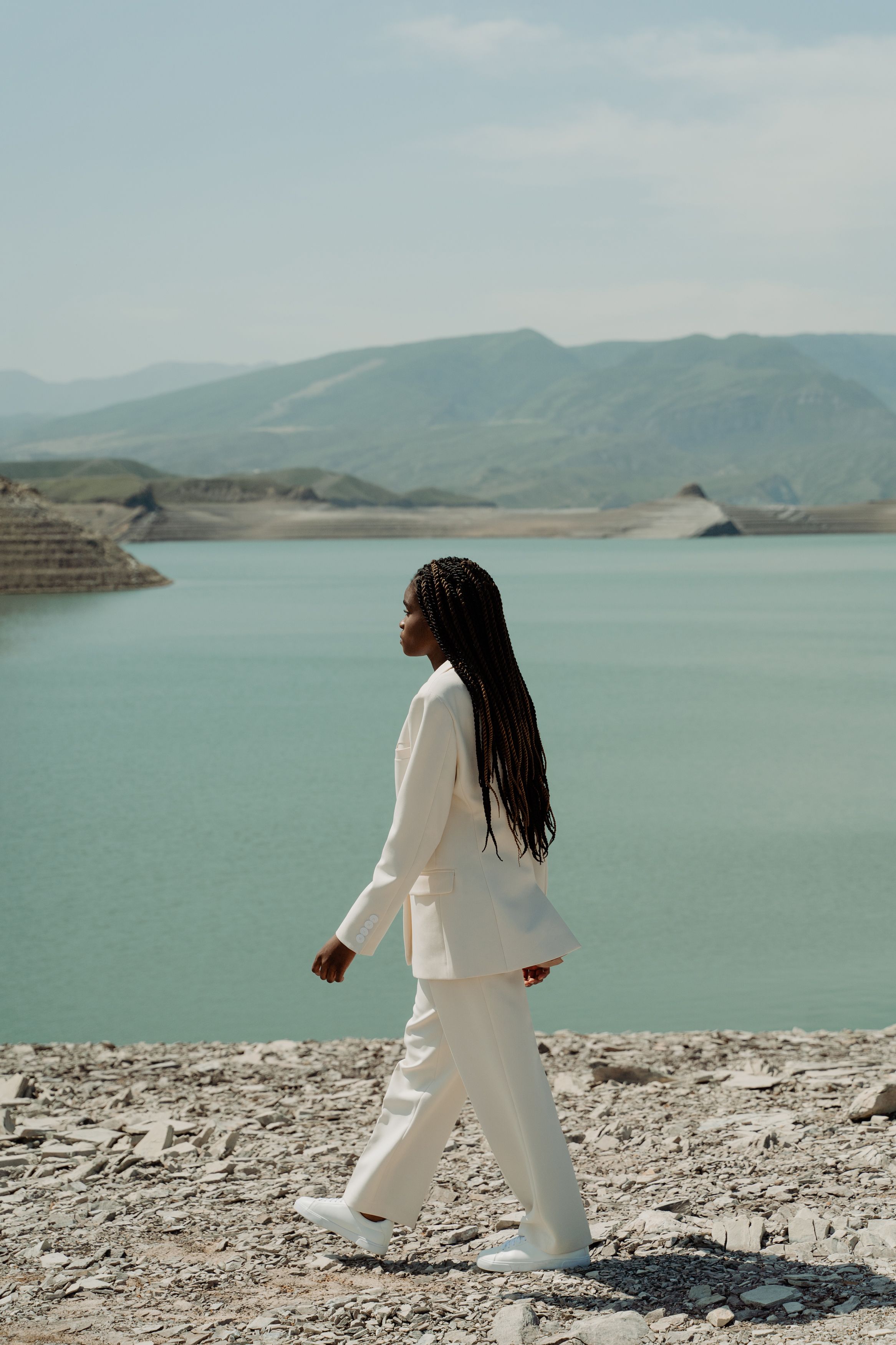 Side view of a fashionable woman in a white suit walking by a tranquil lake with mountains.