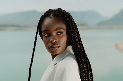 Free Elegant portrait of a fashionable woman in a white suit with afro braids by a serene water backdrop. Stock Photo
