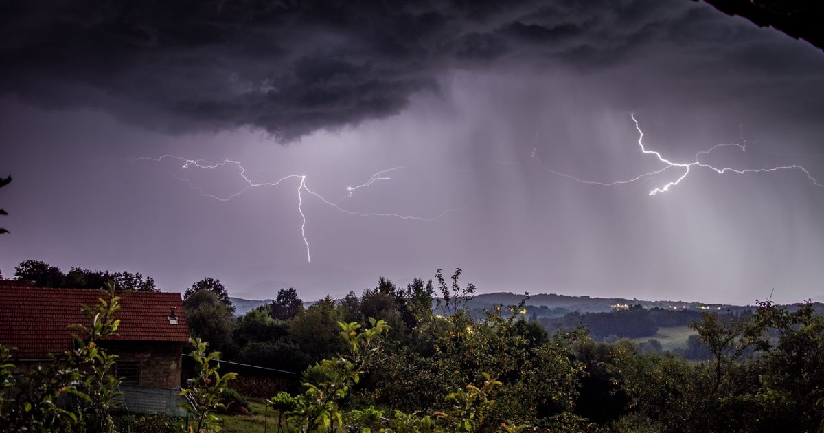 暗い雲の下で何本もの稲妻が走り、手前に家の屋根と木々が見える