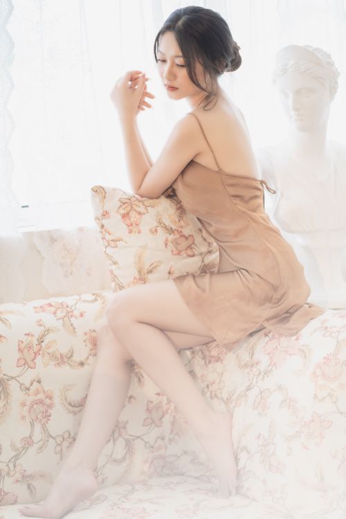 Free A woman in a satin dress sitting on floral couch by a window with soft lighting. Stock Photo