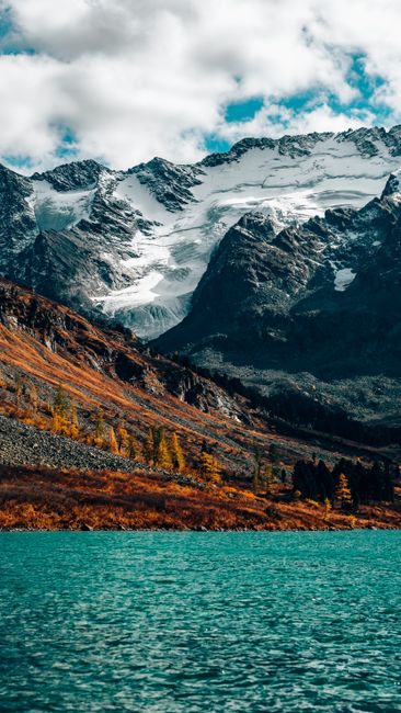 Snowy rocky mountains with a tranquil turquoise lake in the foreground under a partly cloudy sky.