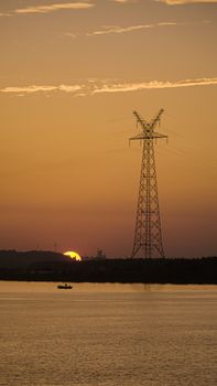 Silhouette of a transmission tower against a vivid sunset sky over a tranquil lake.