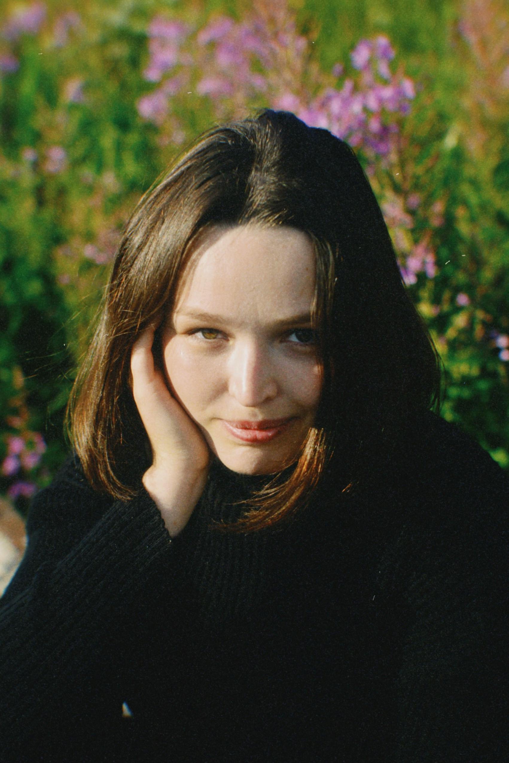 A smiling woman in knitwear poses outdoors with her hand on cheek. Bright floral background.