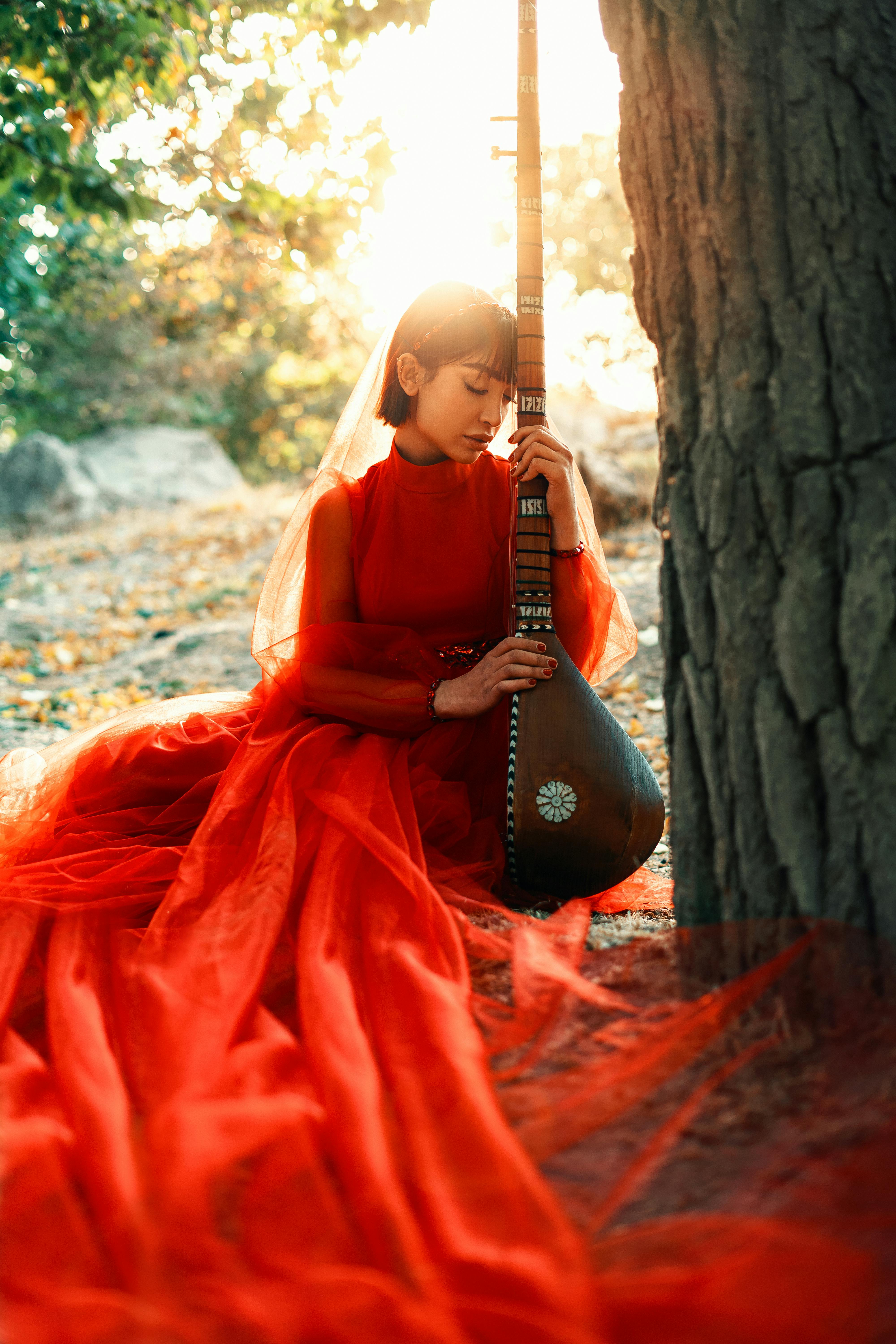 A woman in a red dress elegantly plays a tanbur outdoors, basking in sunlight.