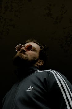 Stylish bearded man in sunglasses and Adidas jacket, gazing upward against a dark background.