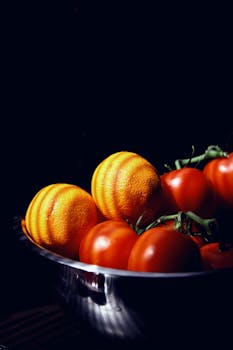Vibrant oranges and ripe tomatoes in a stainless steel bowl against a dark background, showcasing natural freshness.