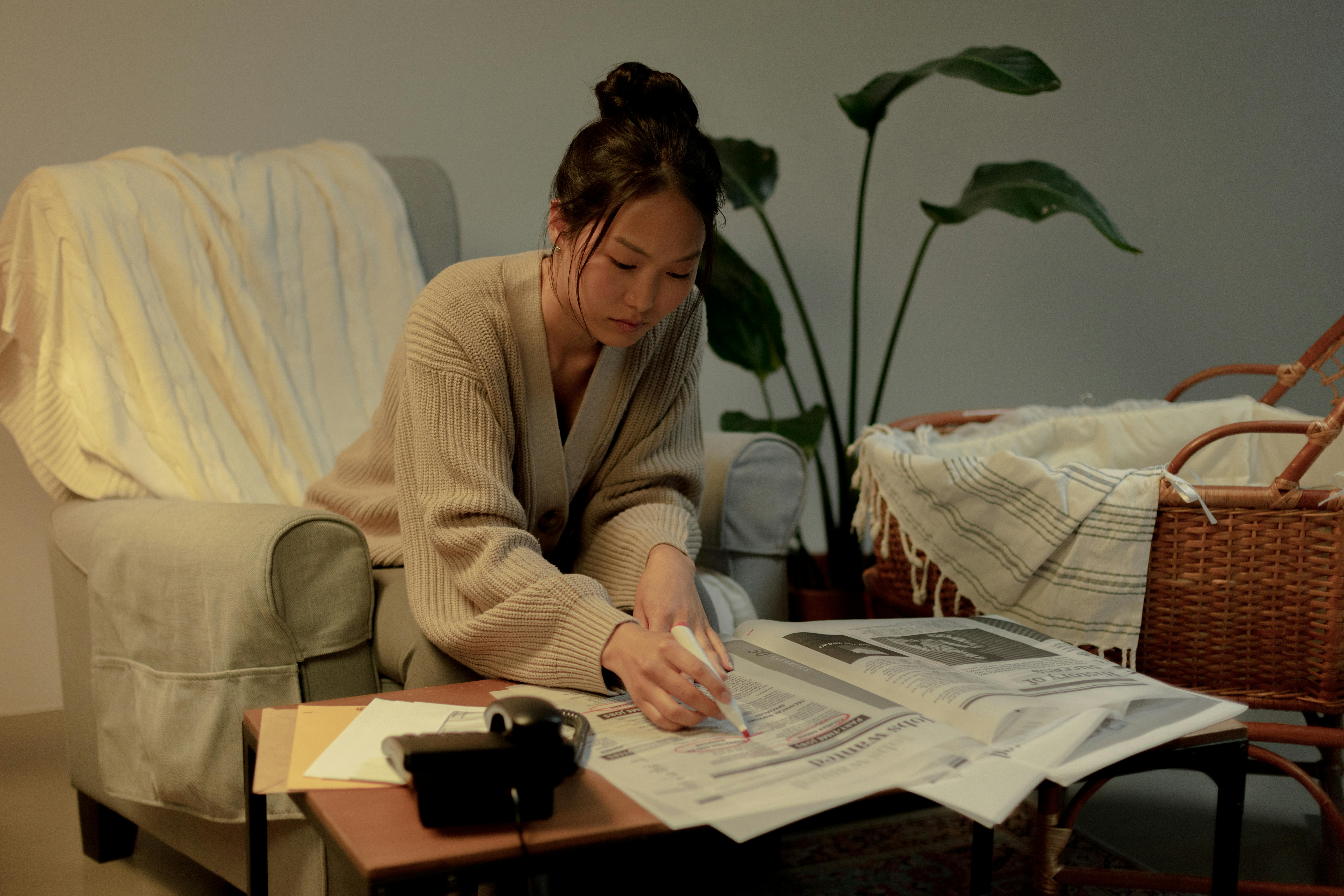 A woman sits in a cozy living room, marking a newspaper with a pen, surrounded by plants and furniture.