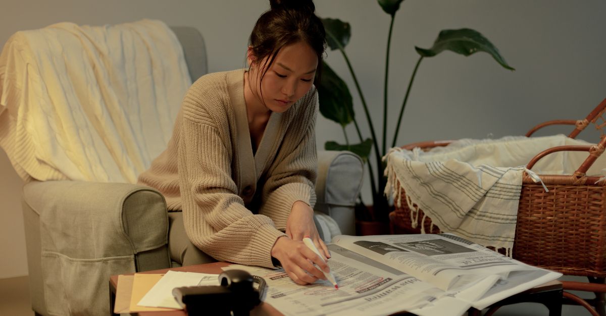 A woman sits in a cozy living room, marking a newspaper with a pen, surrounded by plants and furniture.