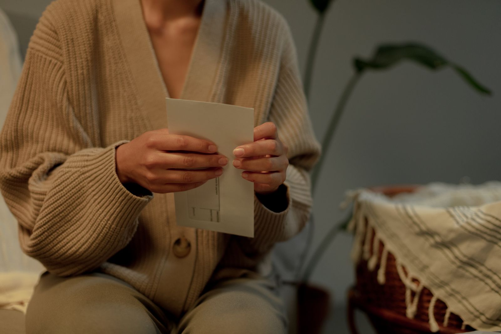 Woman in a beige knit cardigan holding a paper card beside a woven basket and plant