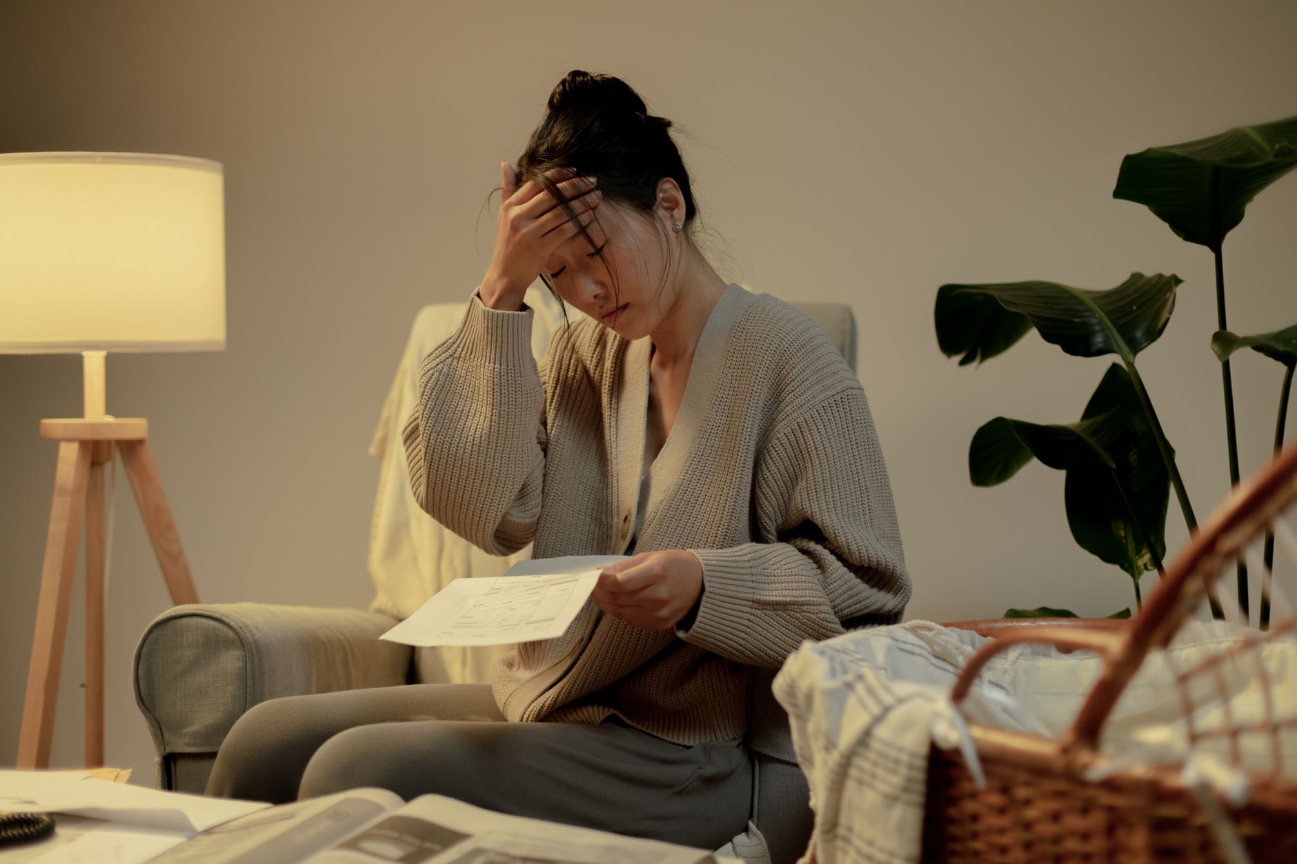 A woman sits in a cozy room, stressed while reading paperwork, hand on head.