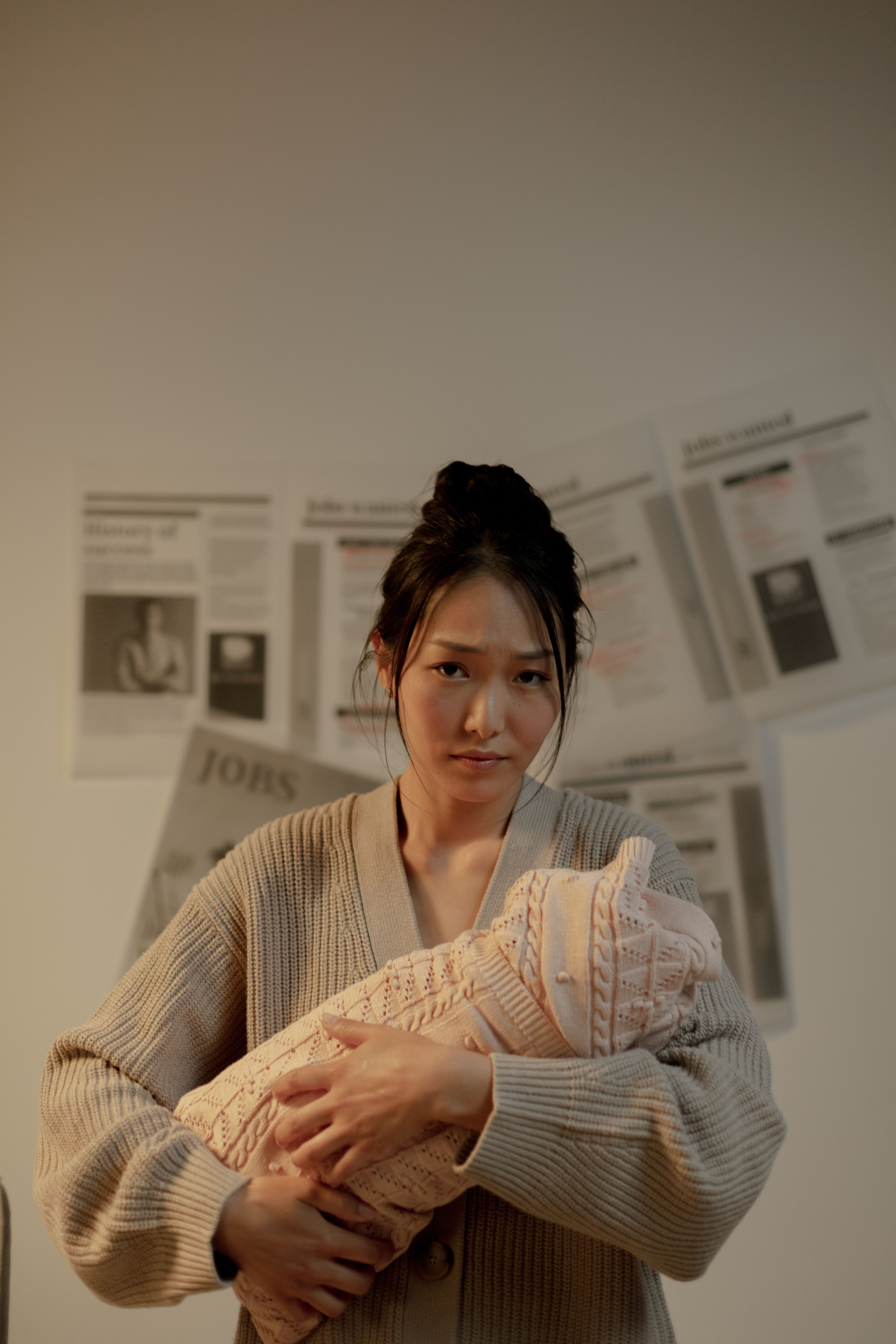 A mother holds her newborn baby indoors, surrounded by newspapers, conveying a thoughtful expression.