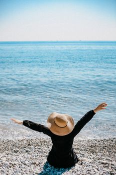 Back view of a woman with arms raised, wearing a hat and sitting by the sea on a pebble beach.