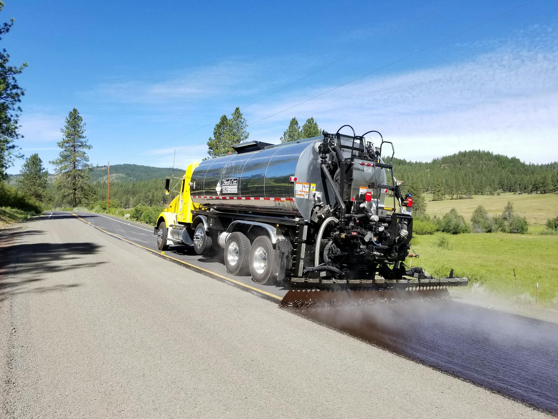 A truck pours asphalt on a sunny highway near lush green fields and trees in Oregon.