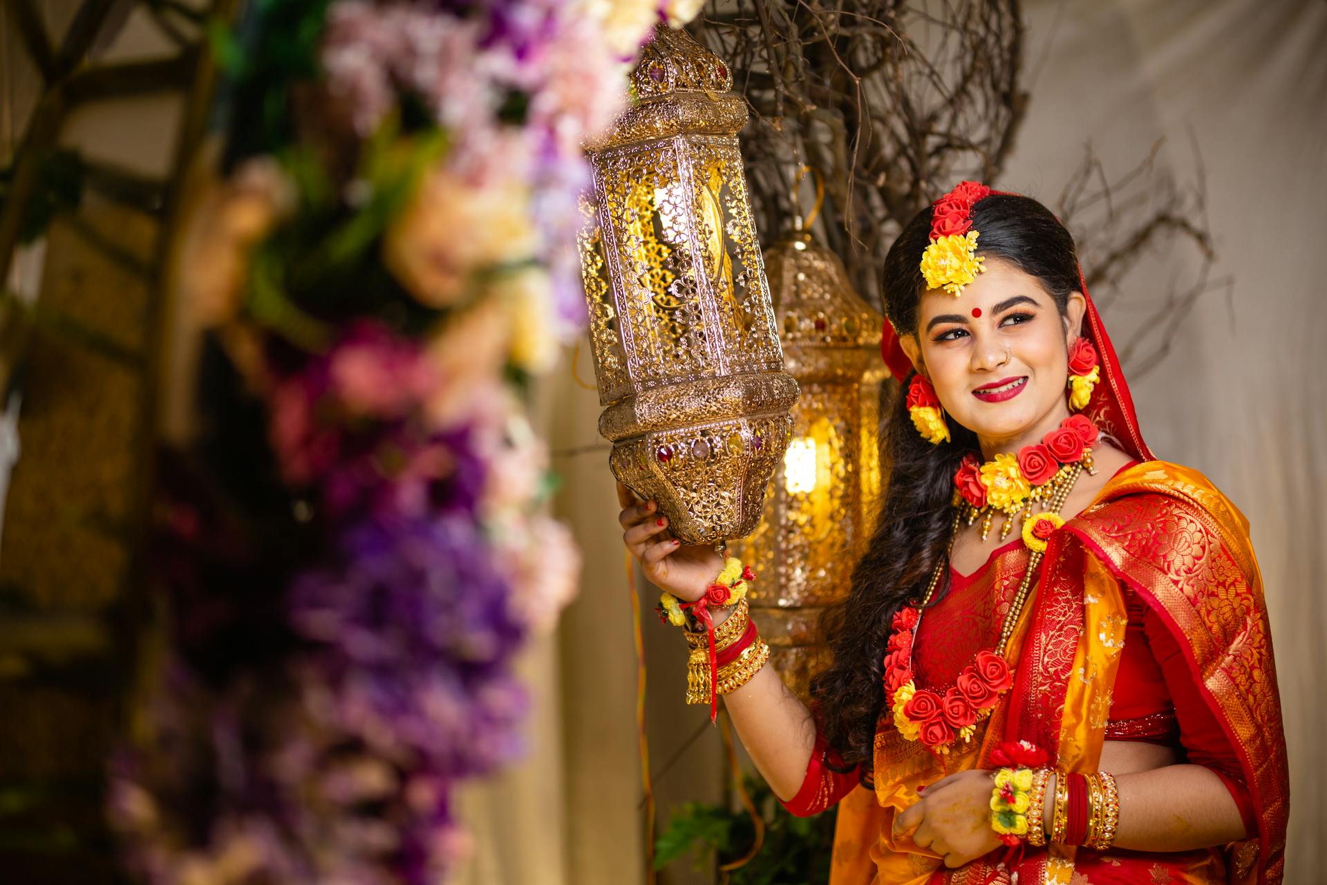 A Bangladeshi bride in vibrant attire adorned with jewelry holding a decorative lamp indoors.