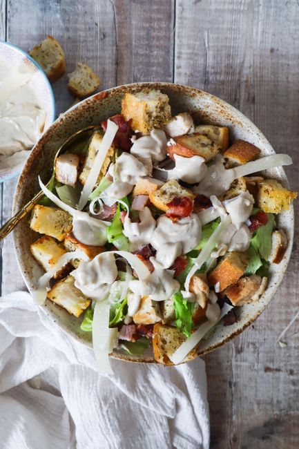 A wooden bowl of creamy chicken salad studded with diced celery and red onion, with a serving spoon resting on the side and a few crackers nearby.