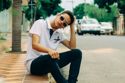 Free Casual young man sitting on a sidewalk by the road wearing sunglasses and a polo shirt. Stock Photo