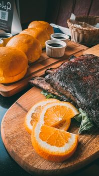 Sliced barbecue ribs with fresh orange slices, buns, and sauces on a wooden board.