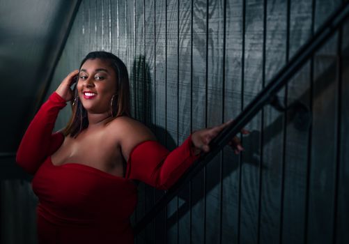 A confident woman in a red dress smiles while leaning on a staircase handrail indoors.