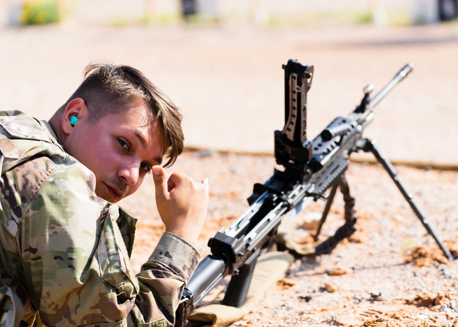 Military personnel in camouflage uniform with a combat rifle on a training field showing a hand gesture.