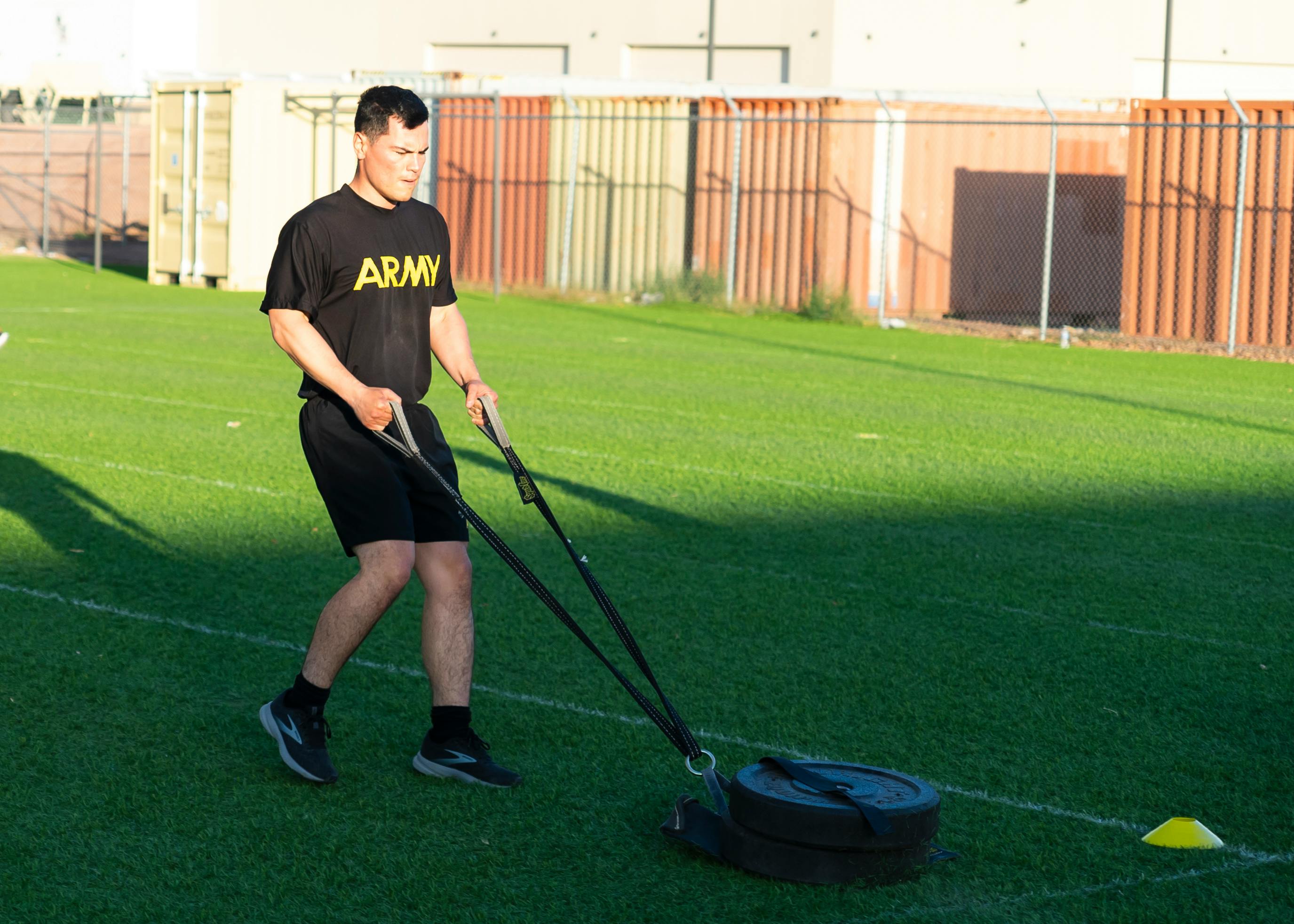 Man pulling a weighted sled during army training on a green field in sunlight.