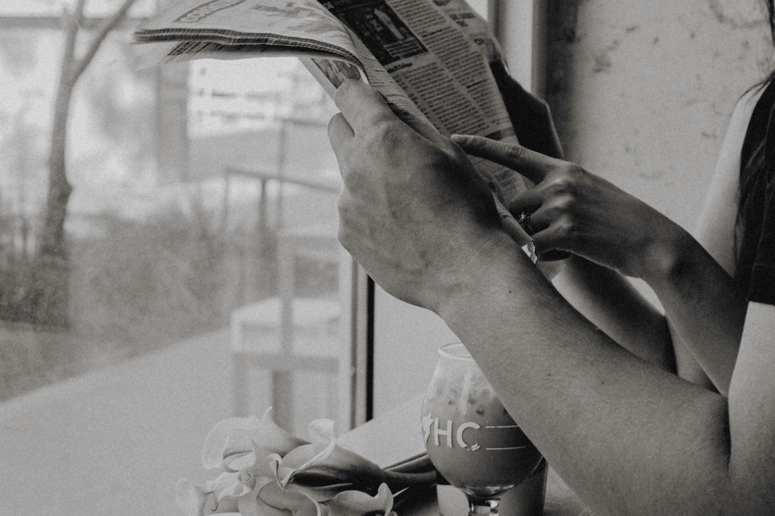 Black and white image of hands holding a newspaper with a drink and flowers on the side.