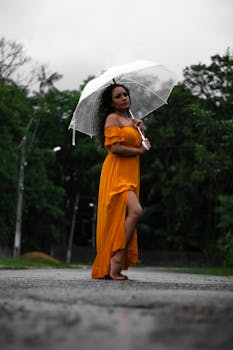 A woman in an orange dress standing barefoot with an umbrella on a street, creating a striking contrast.