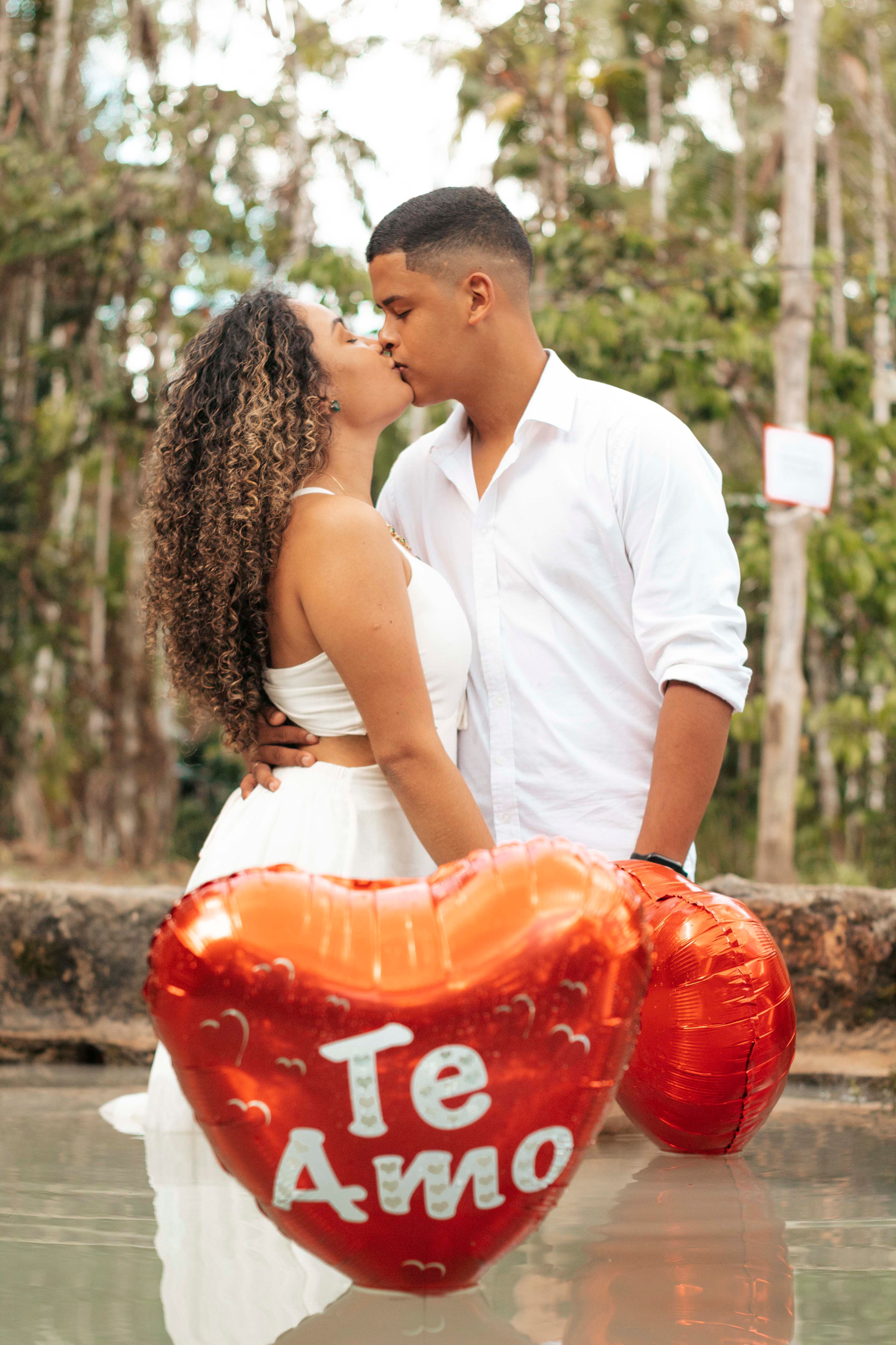 Couple kissing with heart-shaped 'Te Amo' balloons outdoors. Romantic setting with nature background.