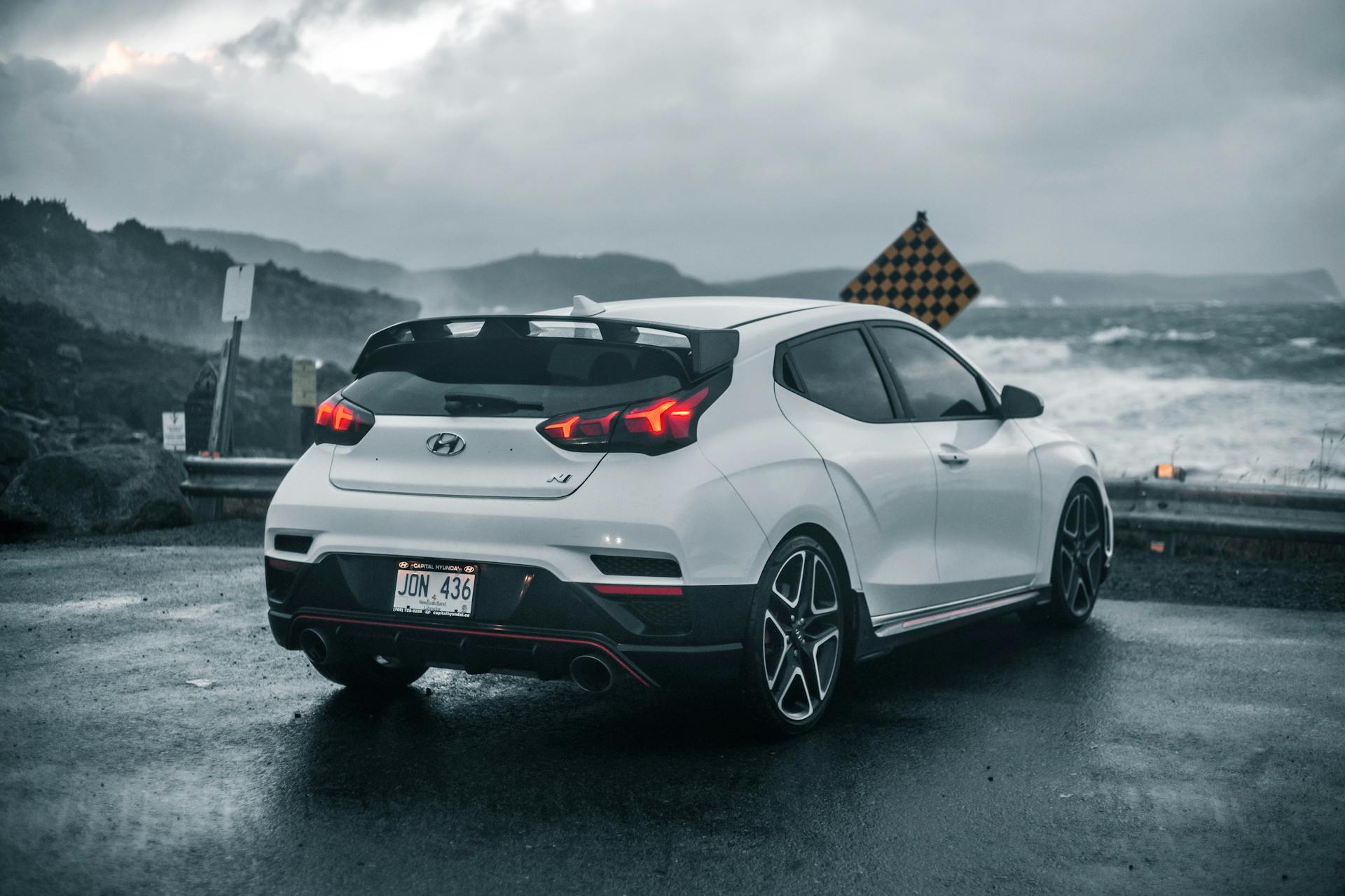 White Hyundai Veloster parked by a wet coastal road under gloomy skies.