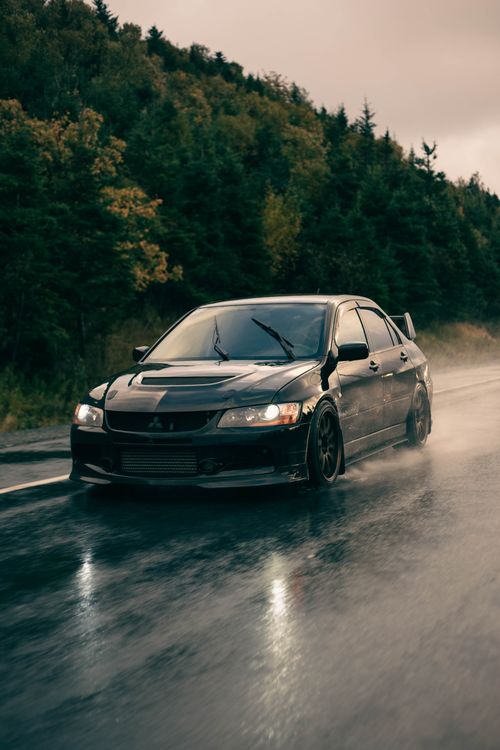 Free A sleek black Mitsubishi Lancer Evolution driving on a wet forest road with a moody atmosphere. Stock Photo