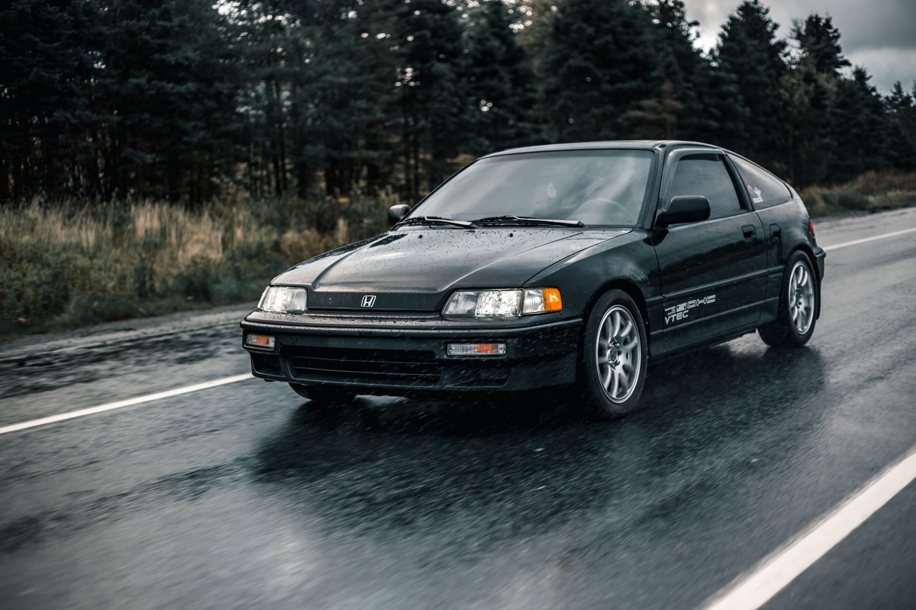 Black Honda hatchback driving on a wet, rural road with forest background.