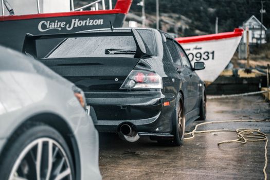 Rear view of a black sports car with spoiler parked near boats at a rainy dockside location.