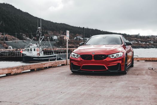 A vibrant red car parked on a dock with a fishing boat and scenic hills in the background.
