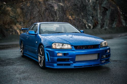 Free A striking blue Nissan Skyline R34 sports car parked on a wet, mountainous road with a misty background. Stock Photo
