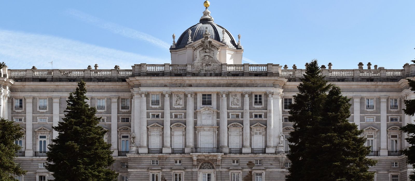 The grand facade of the Royal Palace of Madrid under a clear blue sky, framed by lush trees.