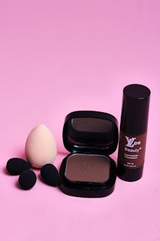 Flat lay of cosmetics including foundation bottle, compact, and sponges on pink background.