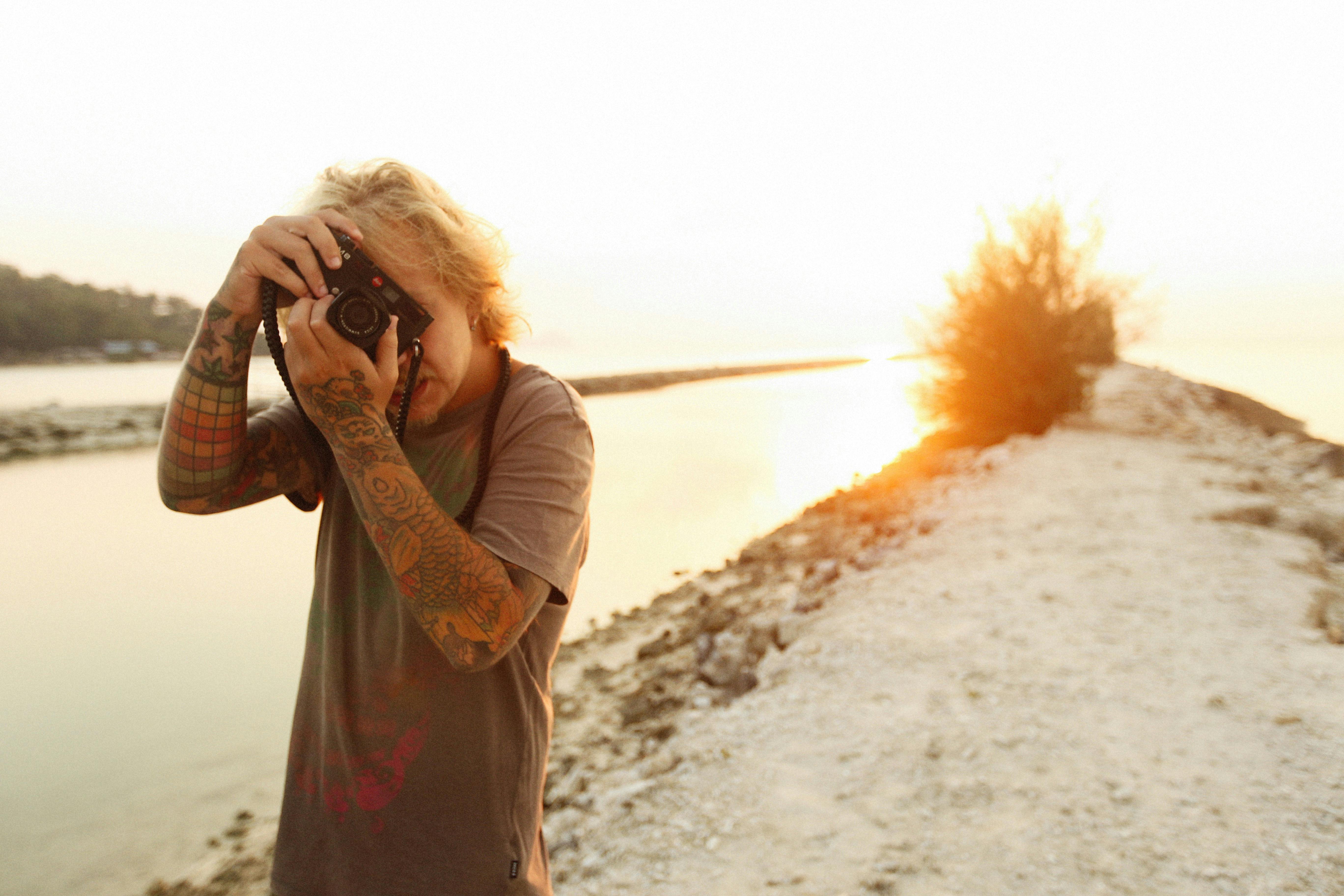 Man with tattoos photographing sunset by the water, showcasing creativity and nature.