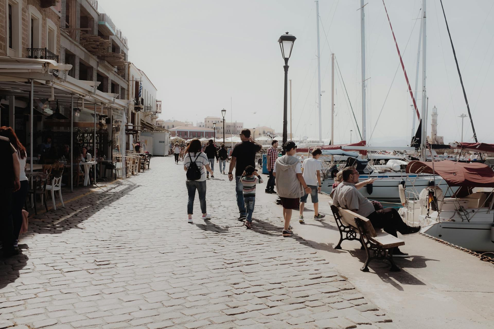Tourists strolling along Chania harbor's cobblestone path, accompanied by docked boats under clear skies.