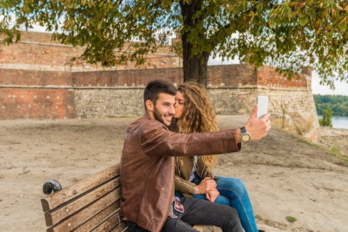Woman And Man Sitting On Brown Wooden Bench Kissing