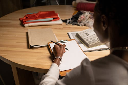 Free Fashion Designer Sketching while Sitting at Desk with Books and Fabric on Stock Photo