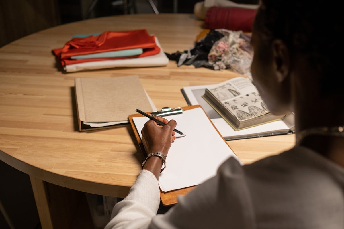Designer di moda seduto al tavolo che disegna accanto a tessuti e libri