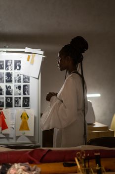 Fashion designer stands in a studio, analyzing designs and photos on a wall, embodying creativity and focus.