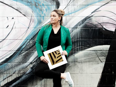 Caucasian woman in green blazer holds artwork against vibrant graffiti wall, outdoors.