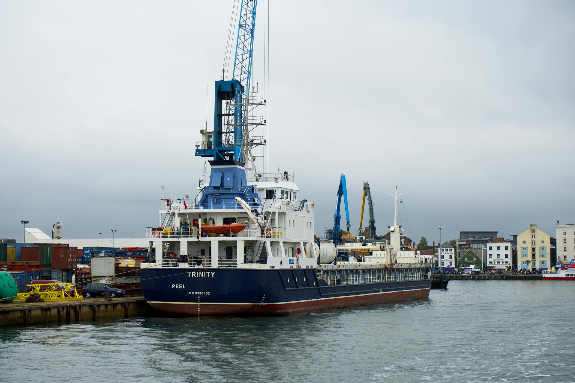 A cargo ship docked at Poole Harbor in England on a cloudy day with cranes and industrial buildings in the background.