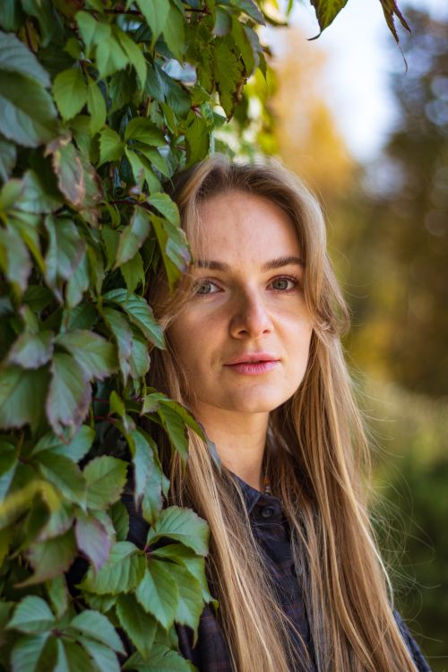 Free Portrait of a blonde woman outdoors, surrounded by lush green leaves. Stock Photo